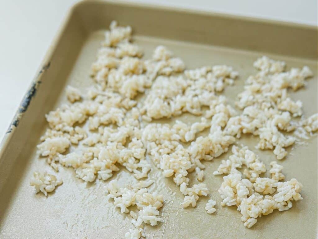 Cooked white rice is spread in clumps across a baking sheet, with some empty spaces visible on the pan’s surface. The rice appears lightly fluffed and scattered.
